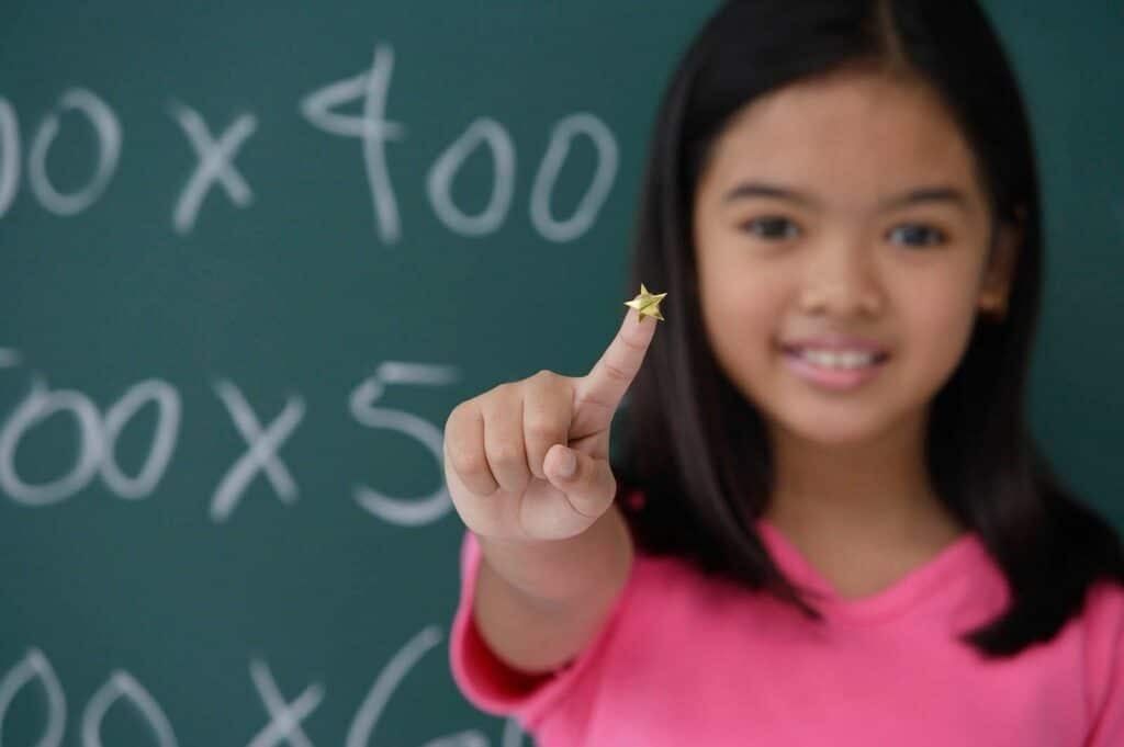 A young girl in front of a chalkboard, smiling and pointing at a star with her index finger