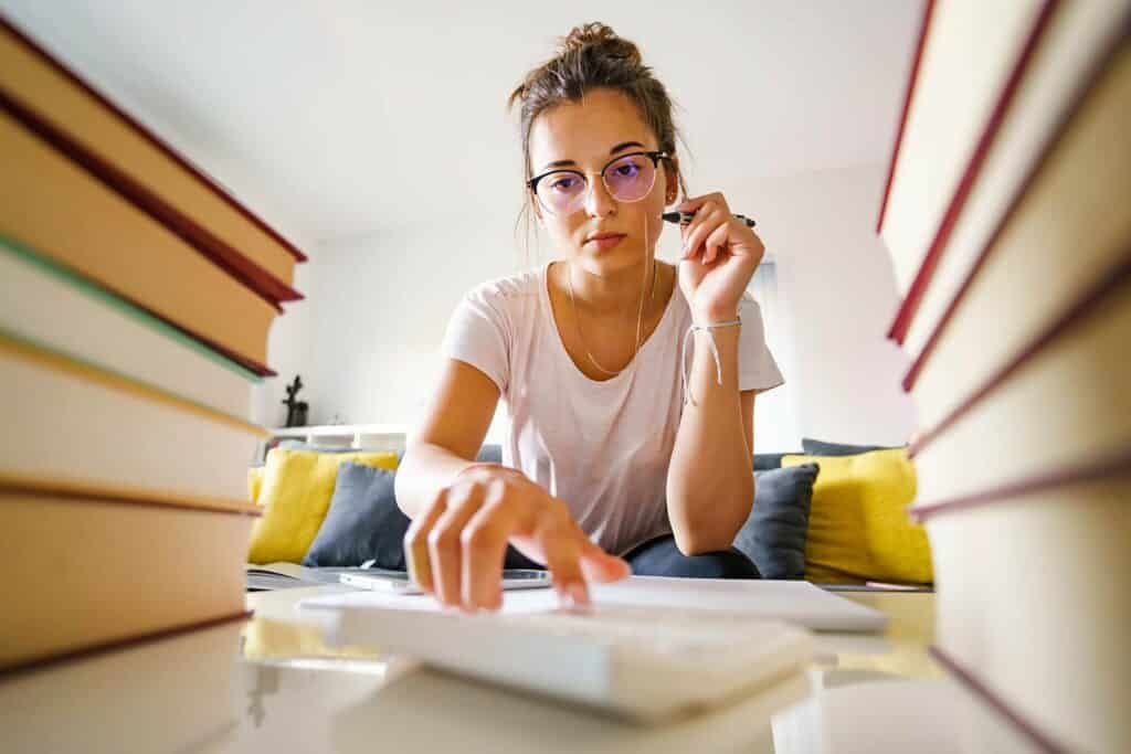 Young caucasian woman female student sitting by the table at home with pile of books reading and writing while study for test - education and learning concept